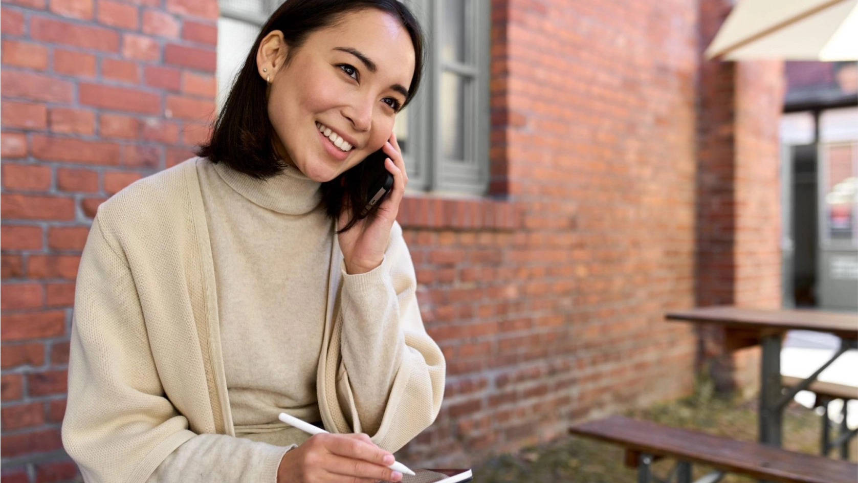 A woman wearing a beige sweater and coat is smiling while talking on her smartphone outside near a brick wall. She holds a stylus and sits at a wooden table, possibly discussing property management banking tasks. Another table and umbrella are in the background.