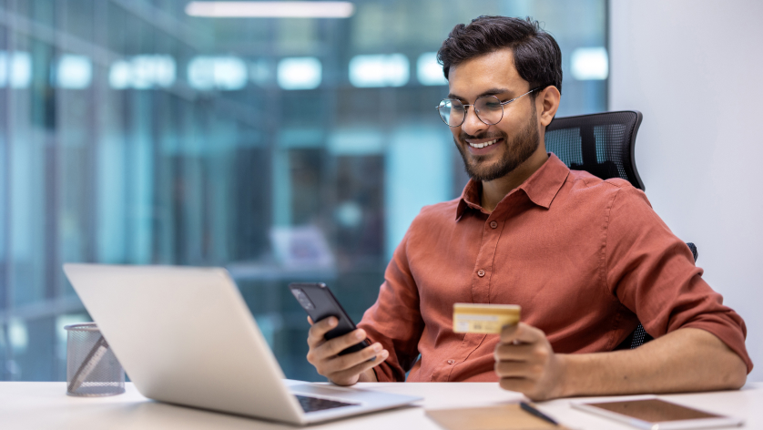 A man sitting at a desk smiles while holding a credit card and looking at his smartphone, possibly checking his escrow bank account. An open laptop and notebook are on the desk, with modern office windows in the background.