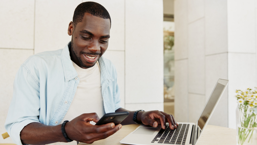 A man sits at a table using a laptop and holding a smartphone, smiling as he manages his ai bank account for real estate. A small bouquet of flowers rests on the table beside him.