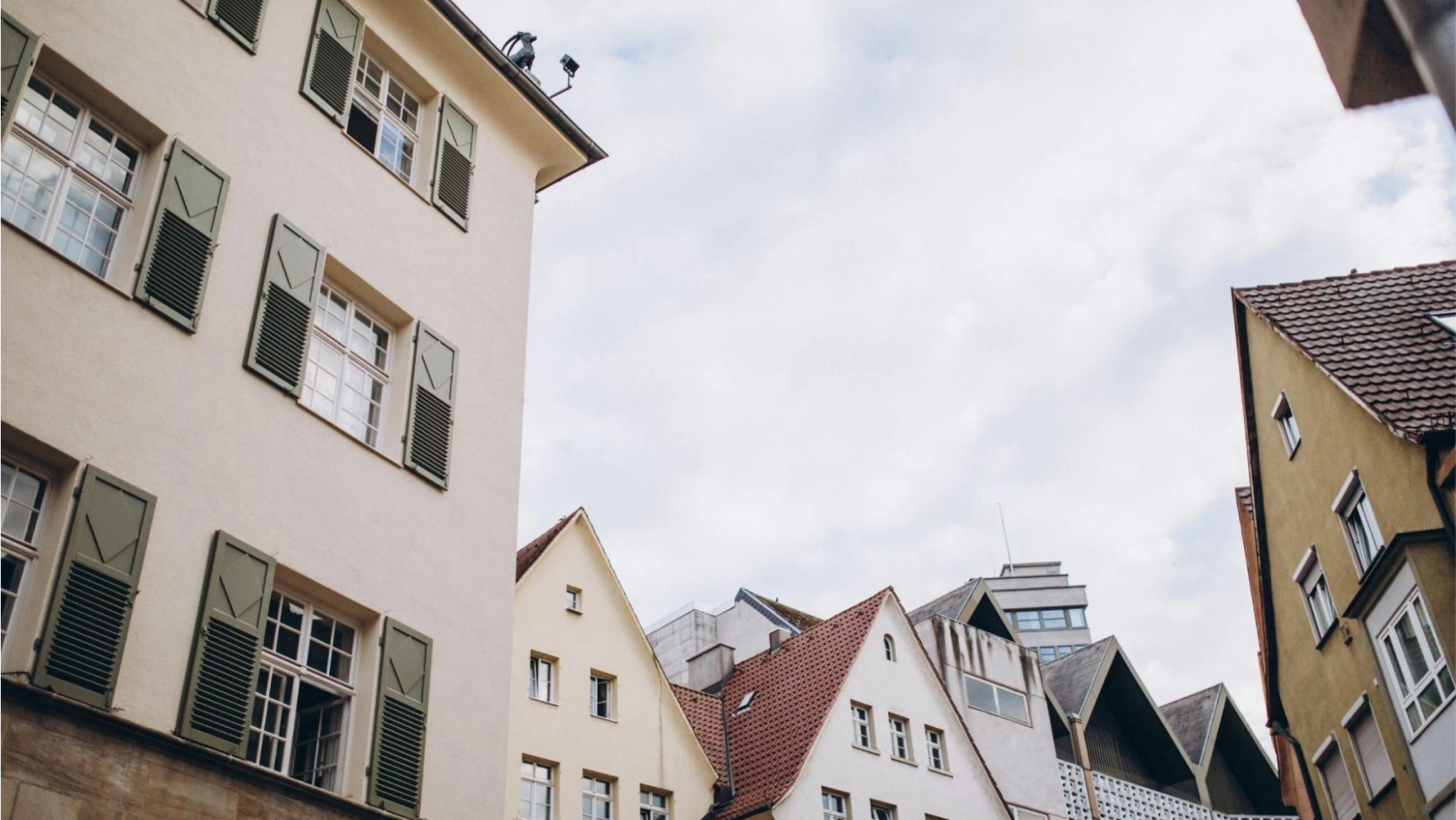 A low-angle view of several traditional European buildings with sloped, red-tiled and gabled roofs, and windows with green shutters—an architectural charm often featured in property management banking portfolios—set against a partly cloudy sky.