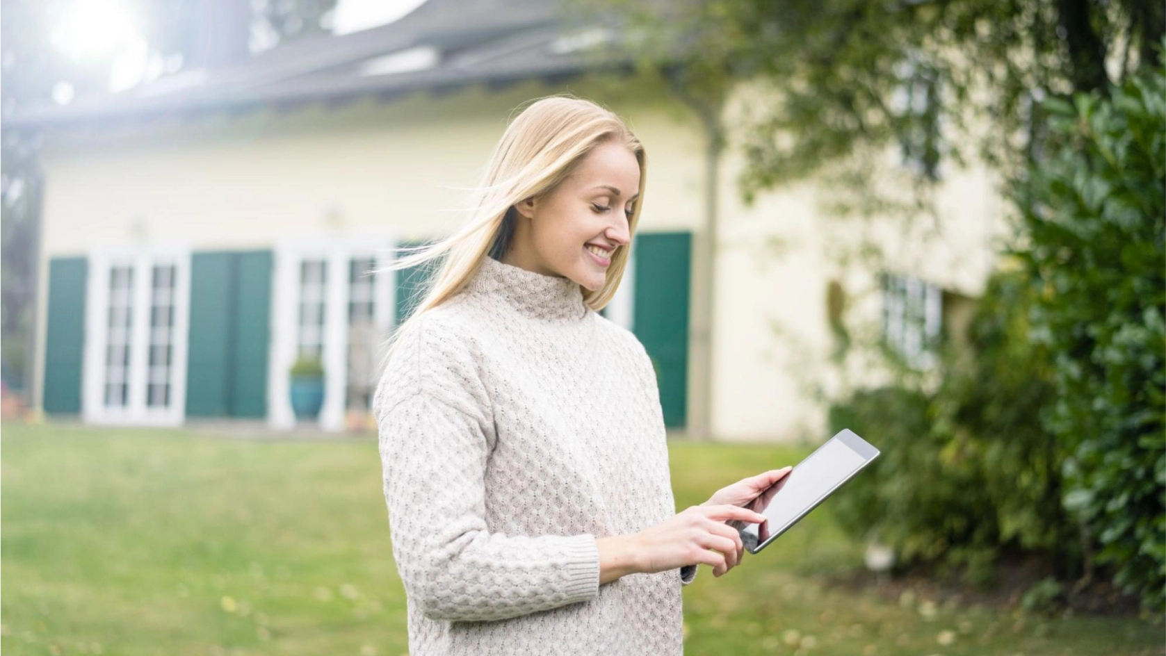 A woman with long blonde hair wearing a light knit sweater stands outside in a green yard, smiling while using her tablet to manage her AI bank account for real estate. A house with white walls and green shutters is visible in the background.