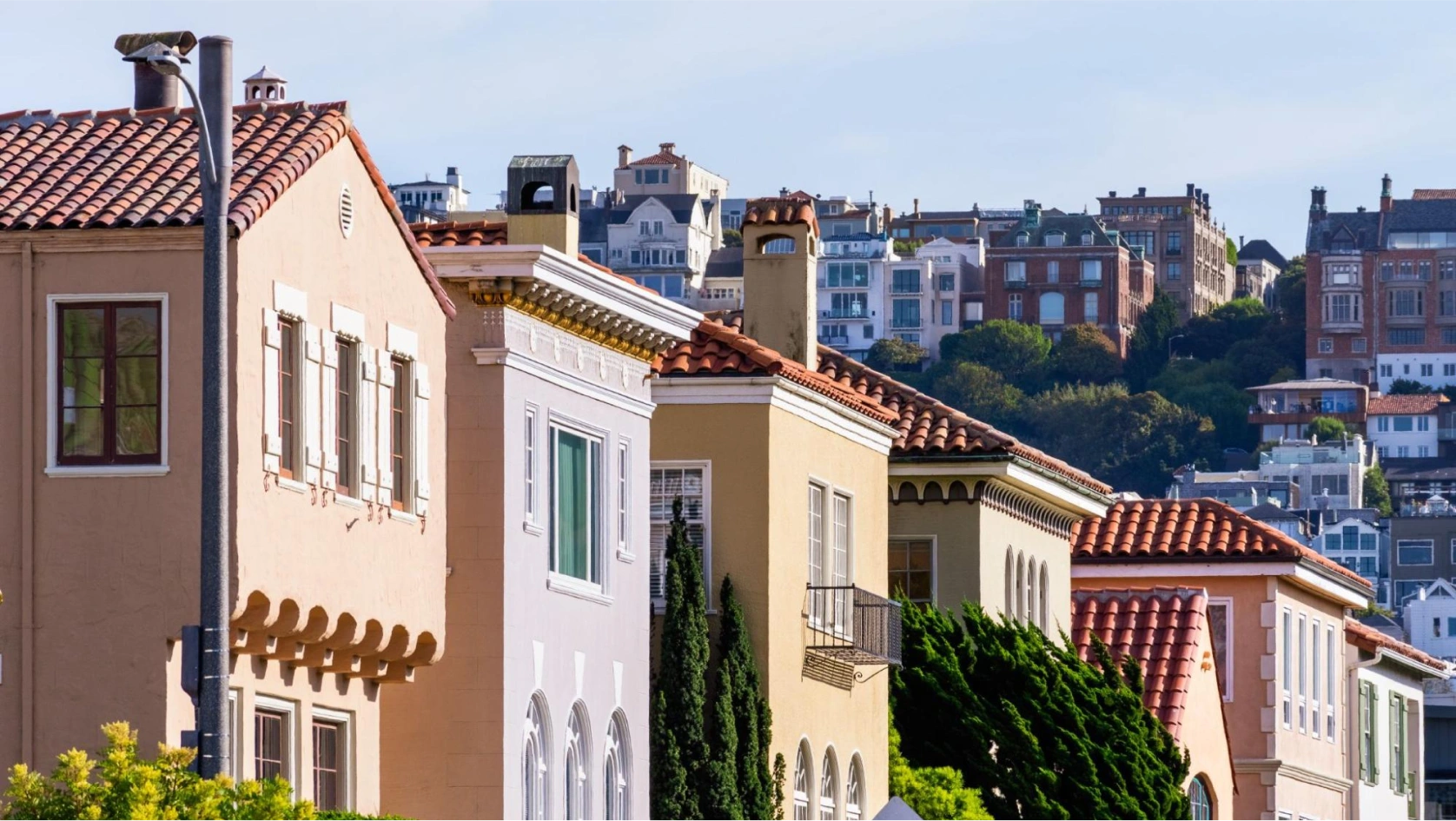 A row of colorful, stucco houses with tiled roofs lines a sunny street, with more homes and greenery visible on a hillside—perfect for exploring options like an AI bank account for real estate investments.