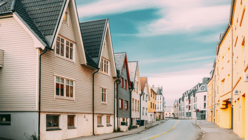 A quiet street lined with colorful wooden houses and buildings, featuring steep gabled roofs under a blue sky with soft clouds. The scene appears calm and empty with no people or cars present.