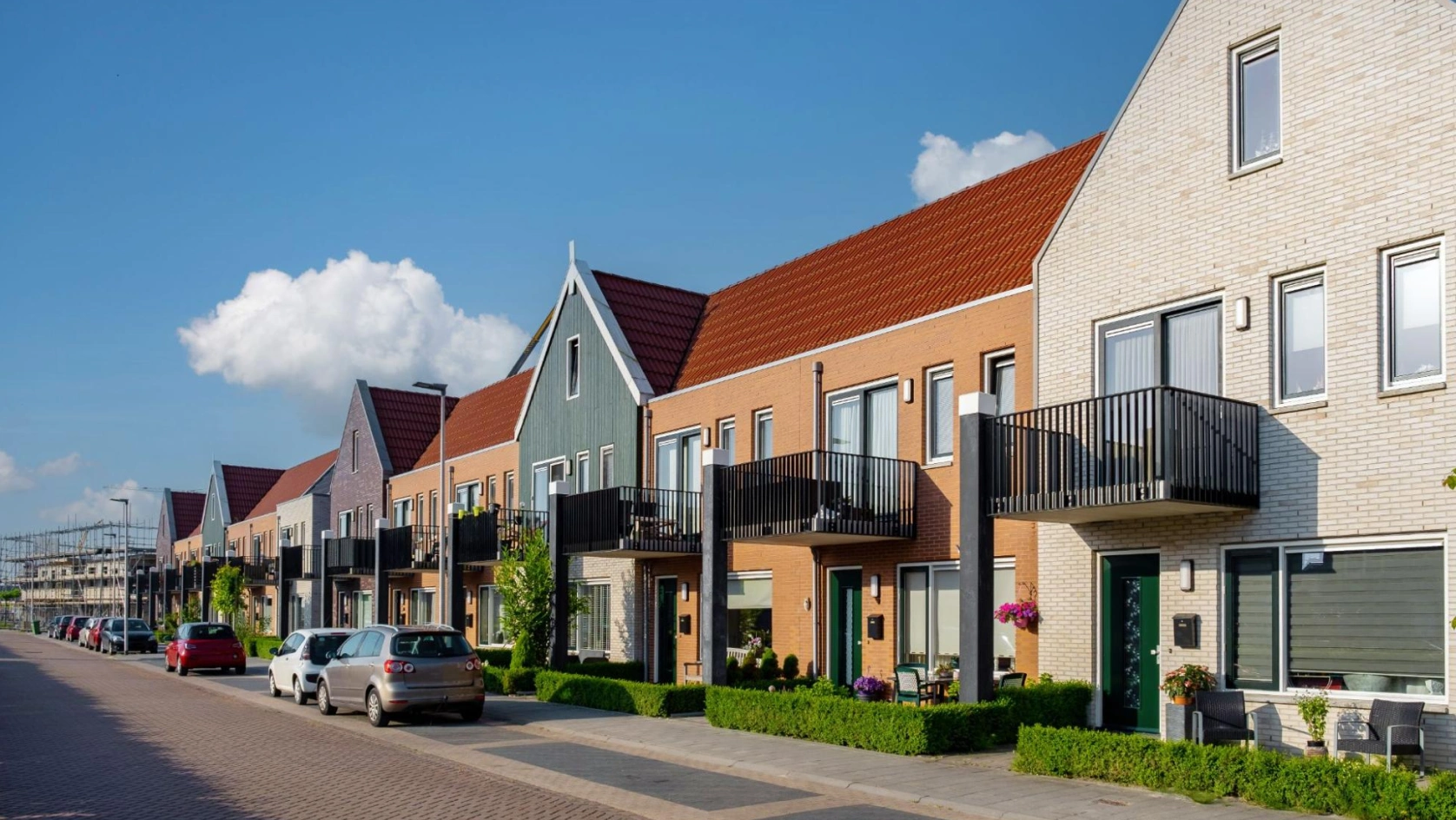 A row of modern, colorful townhouses with gabled roofs and black balconies lines a quiet street—an ideal setting for managing properties with an AI bank account for real estate. Parked cars and trimmed bushes sit under the blue sky.