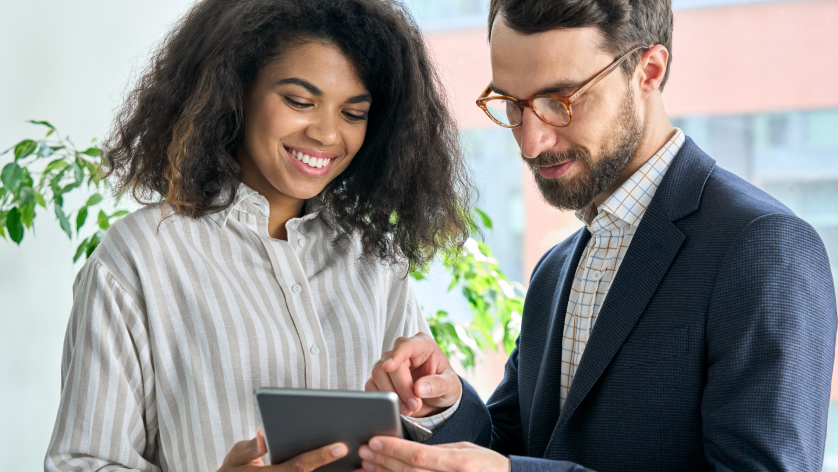 A woman and a man stand together indoors, smiling and looking at a tablet that the man is holding. The woman points at the screen. Both appear engaged in a collaborative discussion.