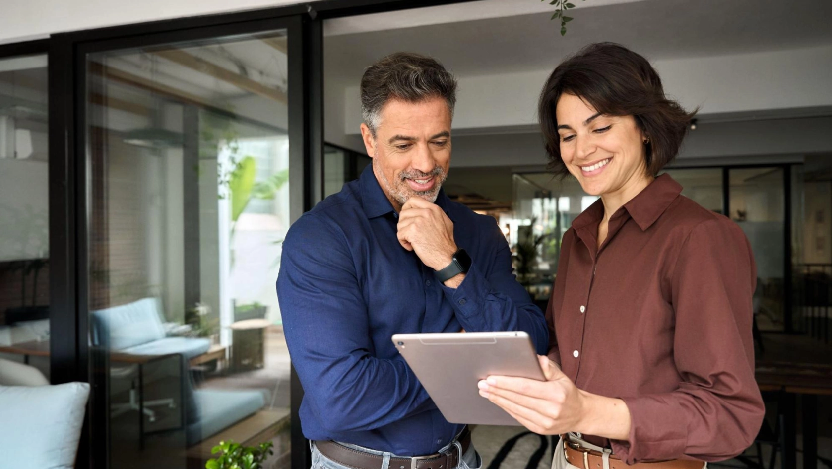 Two smiling adults stand indoors, looking at a tablet together. The man wears a navy shirt and has a thoughtful expression, while the woman in a brown shirt holds the tablet and smiles. Large windows are in the background.
