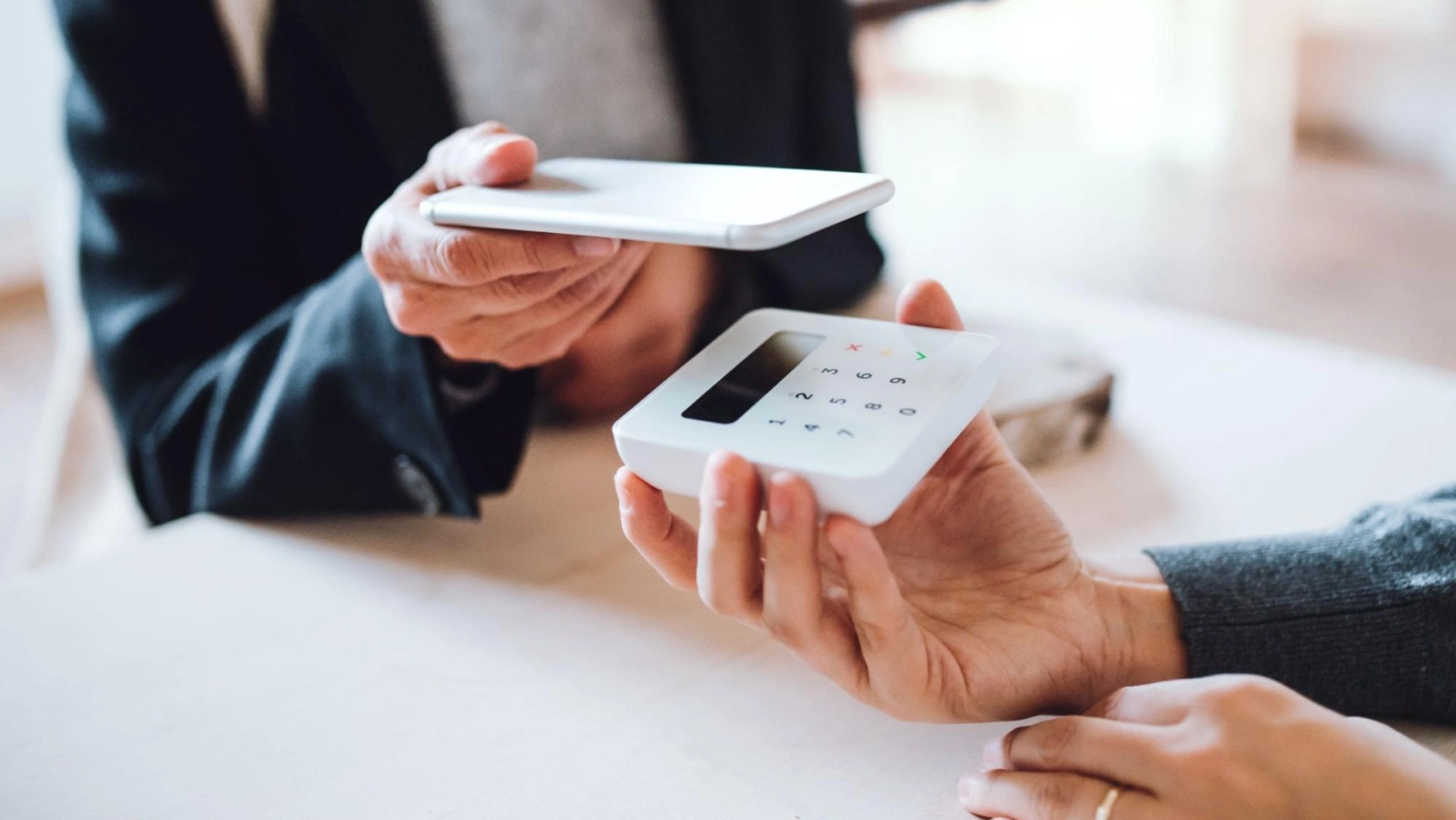Two people make a contactless payment; one hand holds a small card reader while the other holds a smartphone above it, ready to pay. Both individuals are seated at a light-colored table.