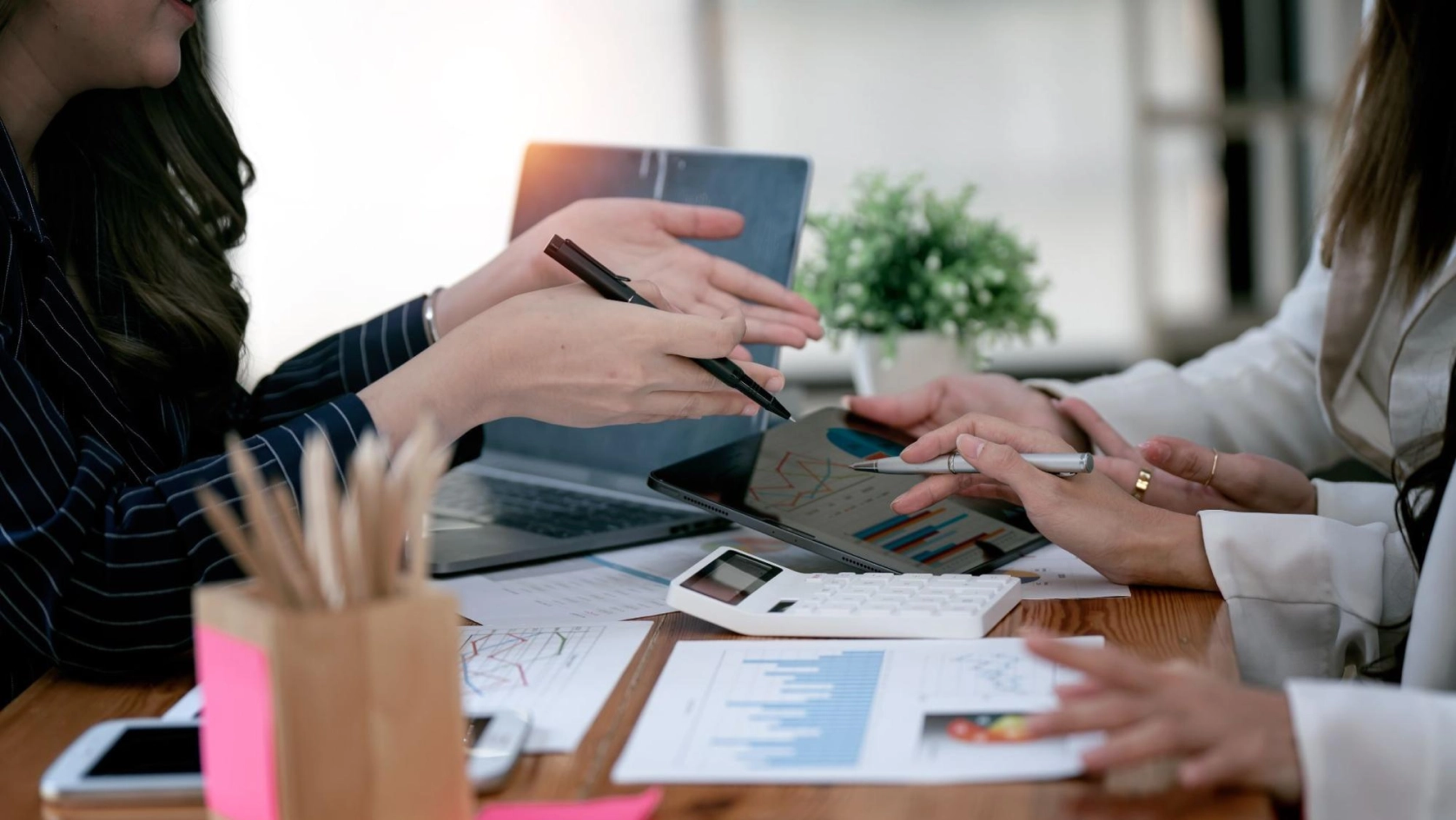 Three people discuss financial charts at a desk with a laptop, tablet, and calculator. Their hands gesture and point at graphs and data, surrounded by office supplies and a small potted plant.