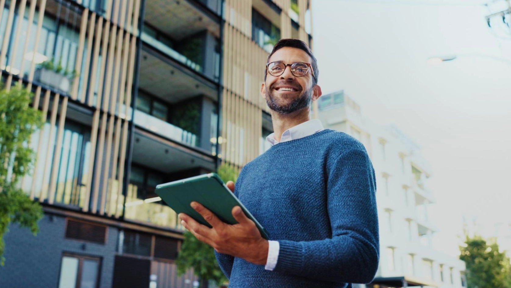A smiling man wearing glasses and a blue sweater stands outdoors in front of modern apartment buildings, holding a tablet in both hands.