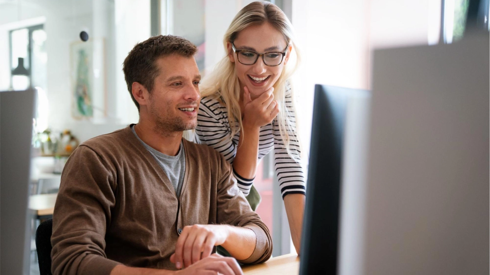 A man sitting at a desk working on a computer, while a woman stands beside him, smiling and looking at the screen. Both appear focused and engaged in a modern office setting.