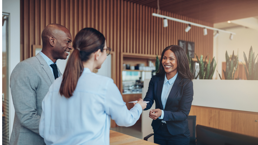 Three professionals in business attire greet each other in a modern office lobby, with one woman smiling and shaking hands with another woman while a man stands beside them, also smiling.