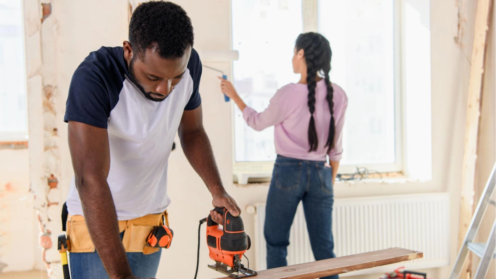 A man uses a jigsaw to cut wood on a workbench while a woman paints a wall near a large window, discussing the importance of separating expenses with a business vs personal bank account during their home renovation.