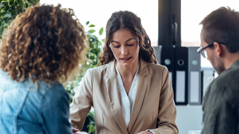 A woman in a beige blazer is talking to two people in an office setting, with plants and binders visible in the background.