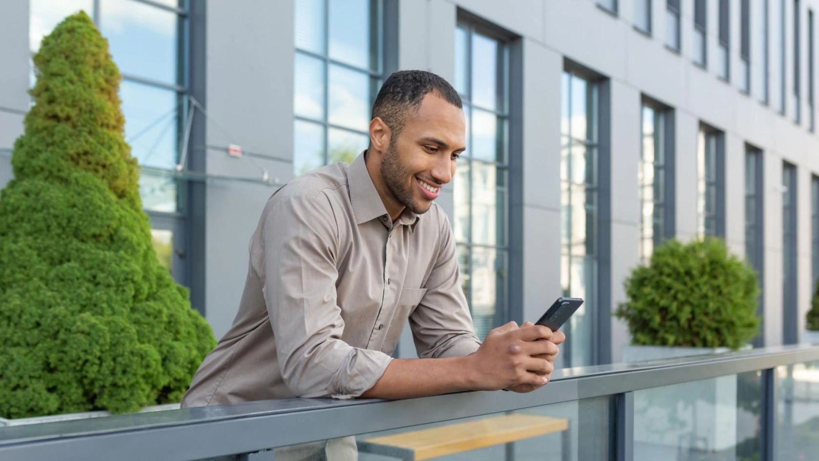 A man in a light brown shirt leans on a glass railing outdoors, smiling while looking at his smartphone. Modern building with large windows and green bushes in the background.