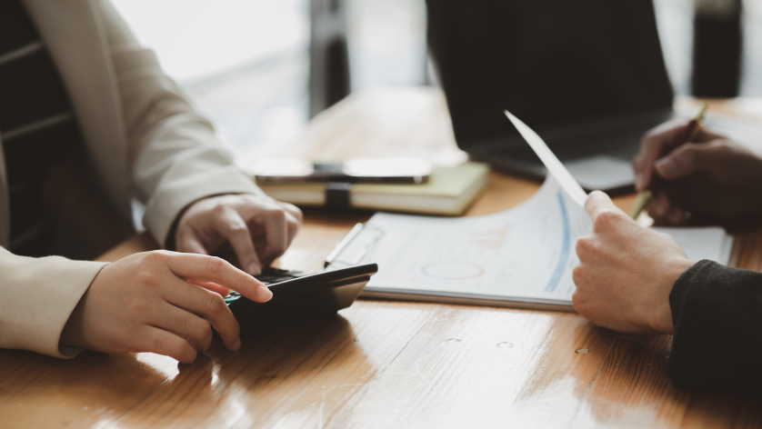 Close-up of two people at a wooden table, one using a calculator while the other reviews an operating reserve account document and writes with a pencil. Papers, a notebook, and a pen are scattered across the surface.