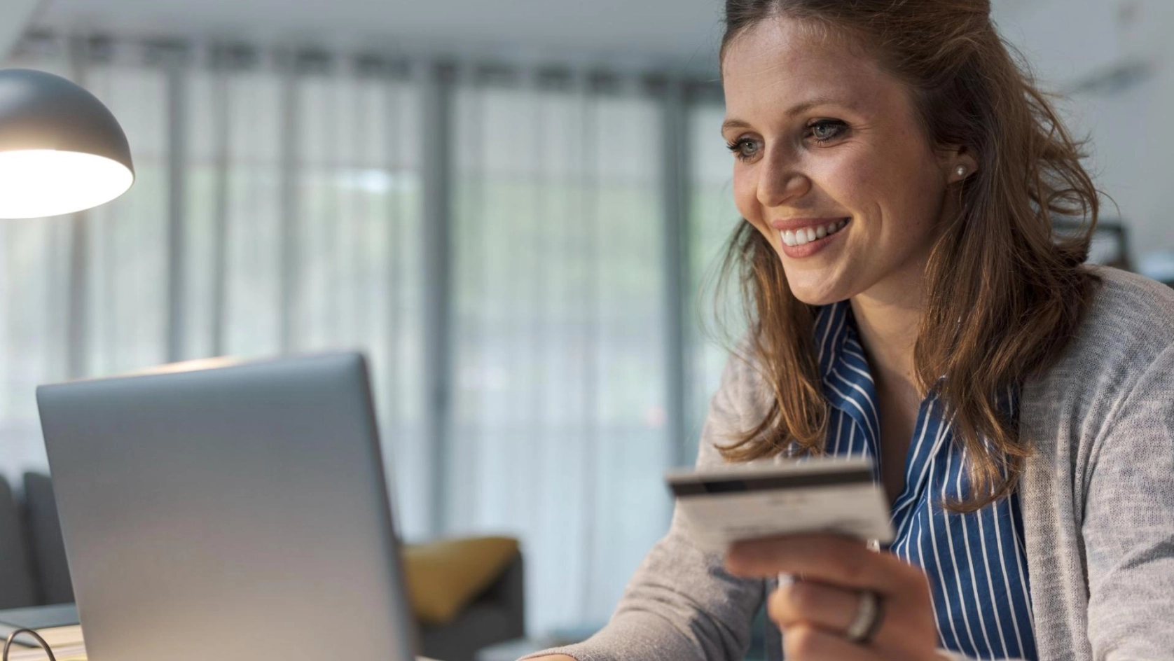 A woman smiles while sitting at a desk and holding a credit card in one hand as she looks at a laptop, suggesting she is shopping or making an online payment at home.