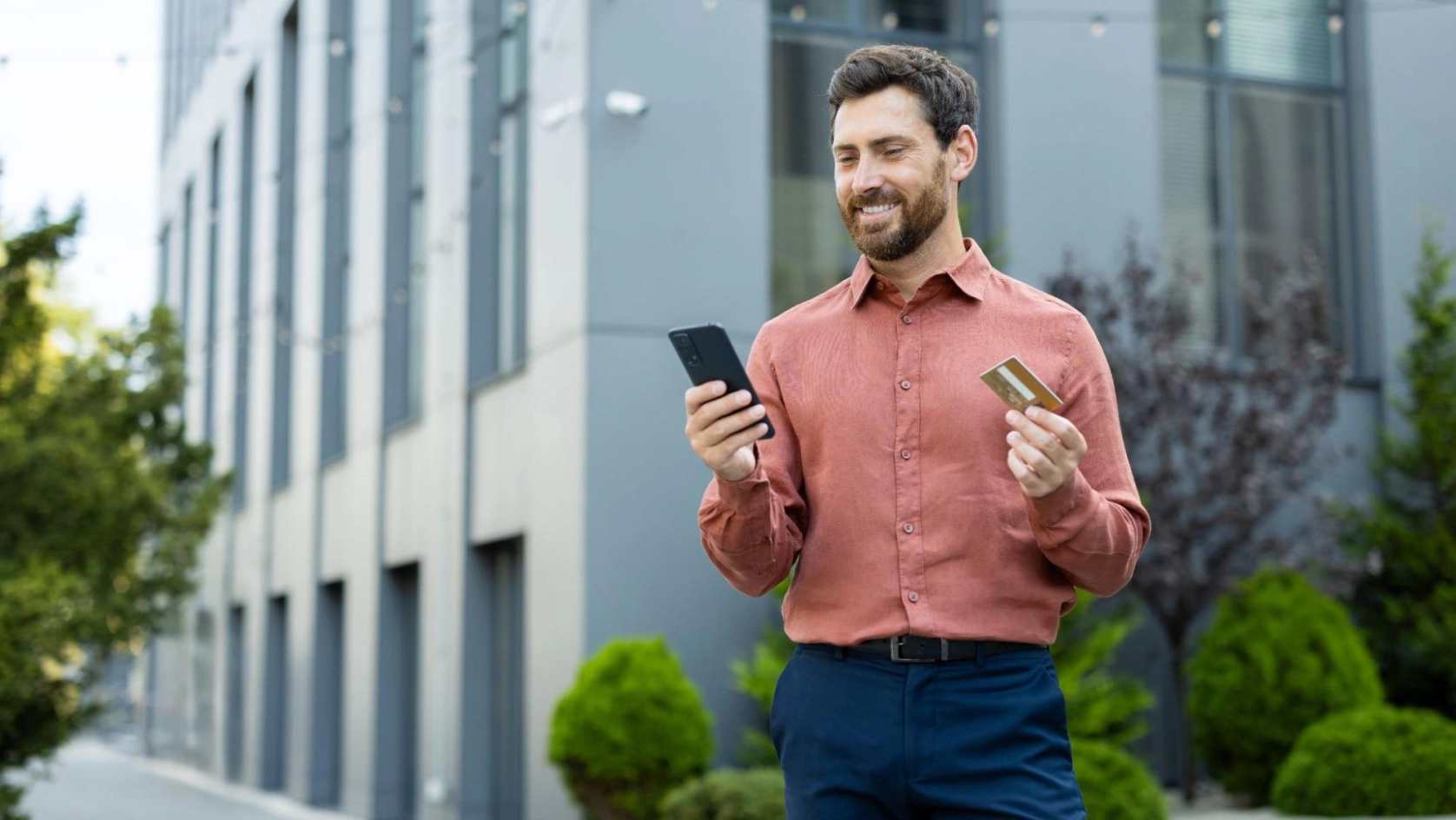 A man in a rust-colored shirt stands outdoors, smiling as he looks at his smartphone and holds a credit card in his other hand, with modern buildings and greenery in the background.