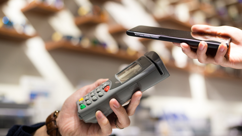 A person holds a payment terminal while another person holds a smartphone close to it, making a contactless payment in a shop. The background is blurred with shelves visible.