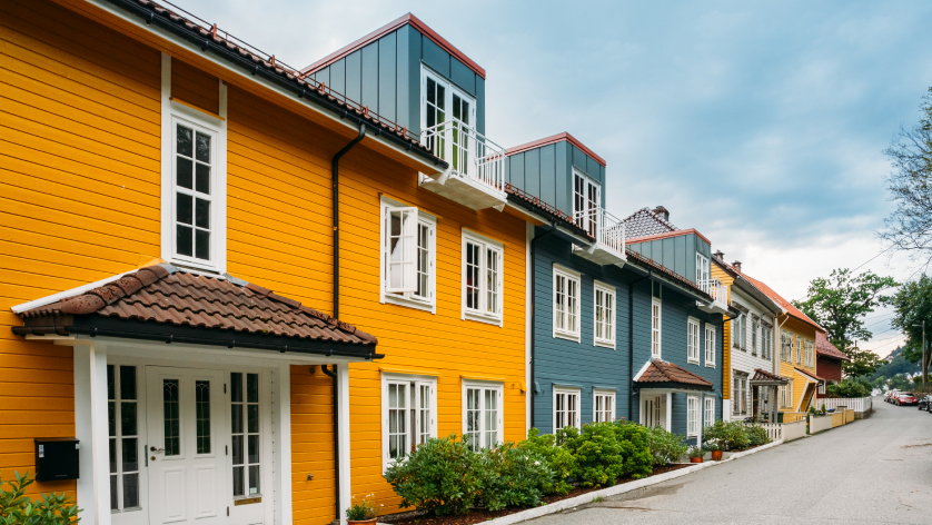 Row of colorful wooden houses, painted yellow, blue, and beige, with white-framed windows and tiled roofs, lined along a quiet street under a cloudy sky.