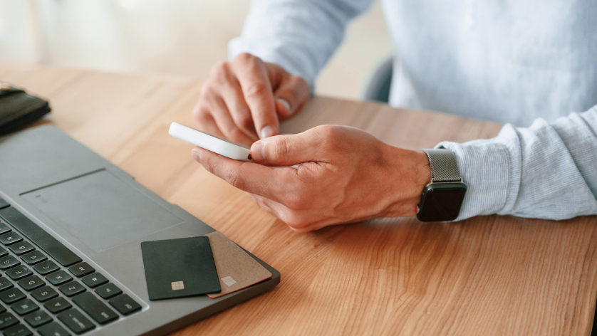 A person wearing a smartwatch uses a smartphone at a desk with a laptop and two credit cards nearby. The focus is on their hands and the devices.
