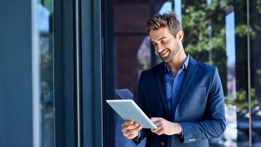 A smiling man in a blue suit stands outside by a glass building, using a tablet. Sunlight and trees are reflected in the windows behind him.
