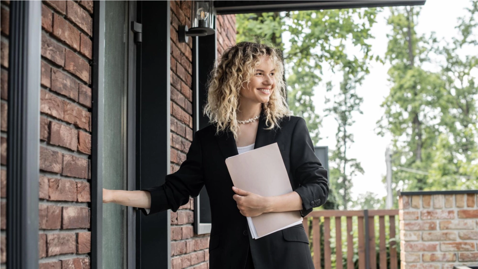 A person with curly blonde hair, wearing a black blazer and holding folders, smiles while standing at the door of a brick building, about to knock or ring the doorbell. Trees and a wooden fence are visible in the background.