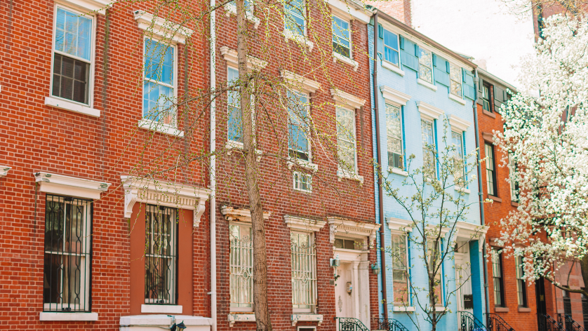 A row of colorful townhouse buildings, mostly red brick with one light blue, stand along a city street lined with blooming trees in spring.