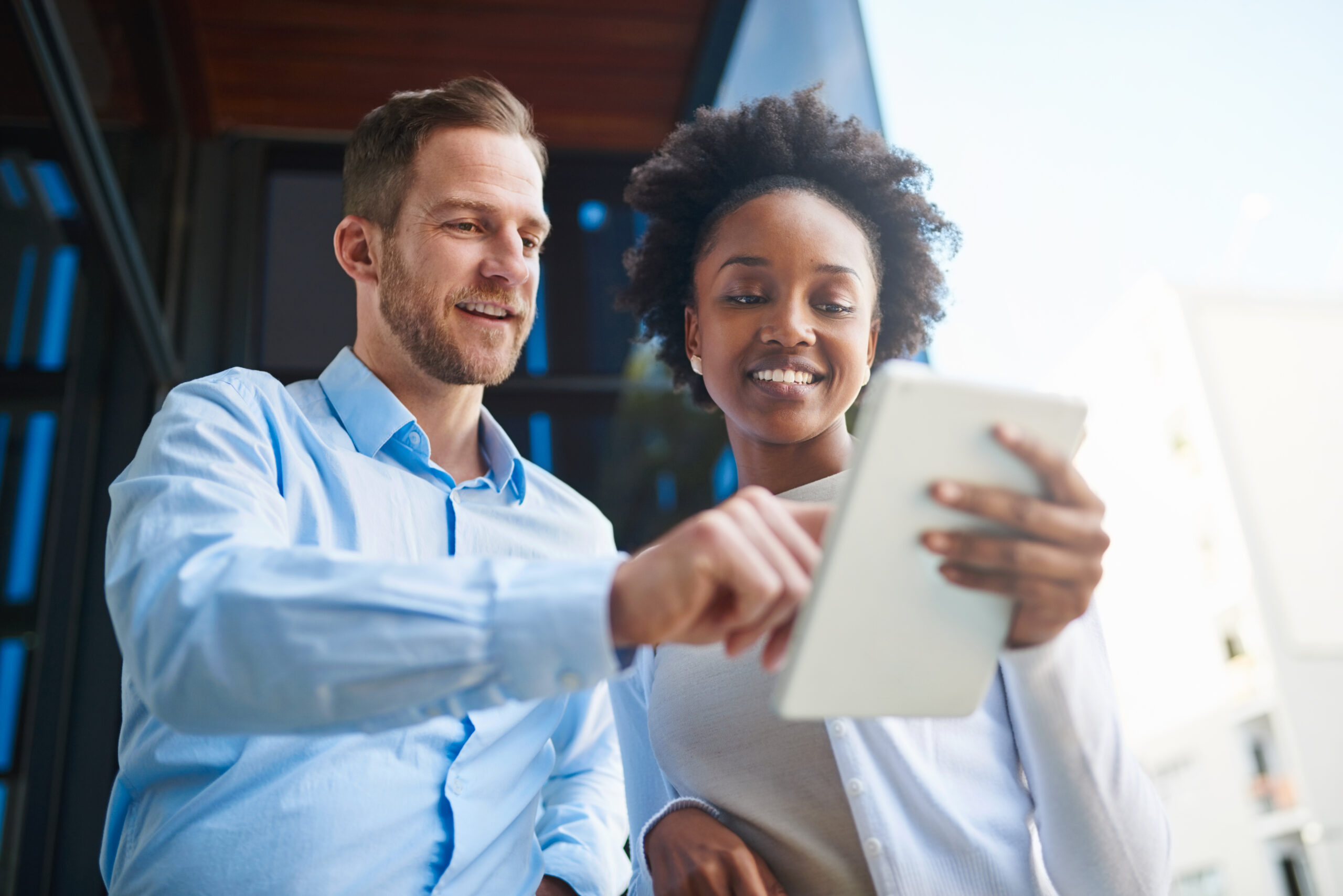 Two people, a man and a woman, stand outside a building smiling while looking at and interacting with a tablet together.