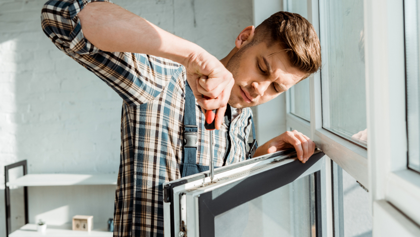 A man in a plaid shirt uses a screwdriver to fix or install a window frame, focusing intently—much like weighing options between a business vs personal bank account—in a brightly lit room.