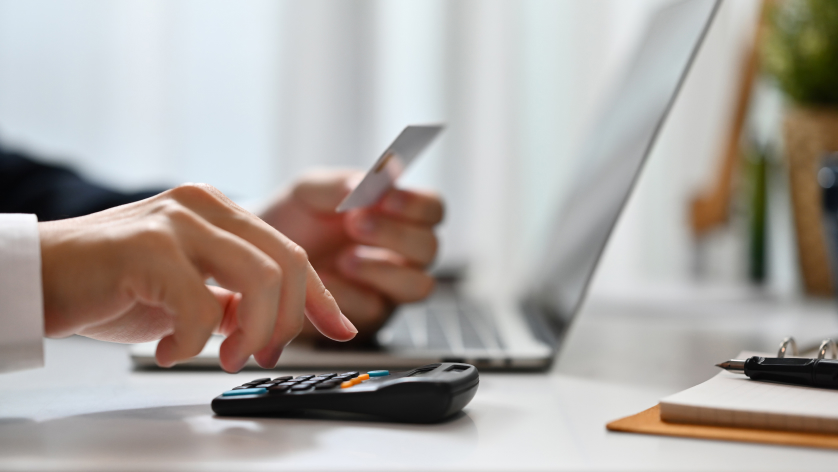 A person uses a calculator while holding a credit card and working on a laptop at a desk, suggesting online shopping or financial planning.