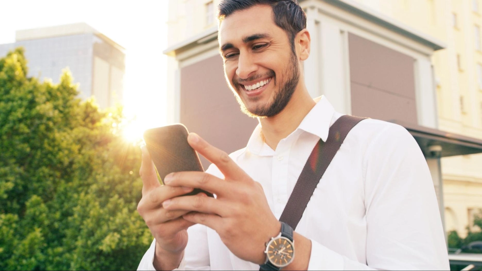 A smiling man in a white shirt stands outdoors in sunlight, looking at his smartphone. He has a shoulder bag and wears a wristwatch, with buildings and green trees in the background.
